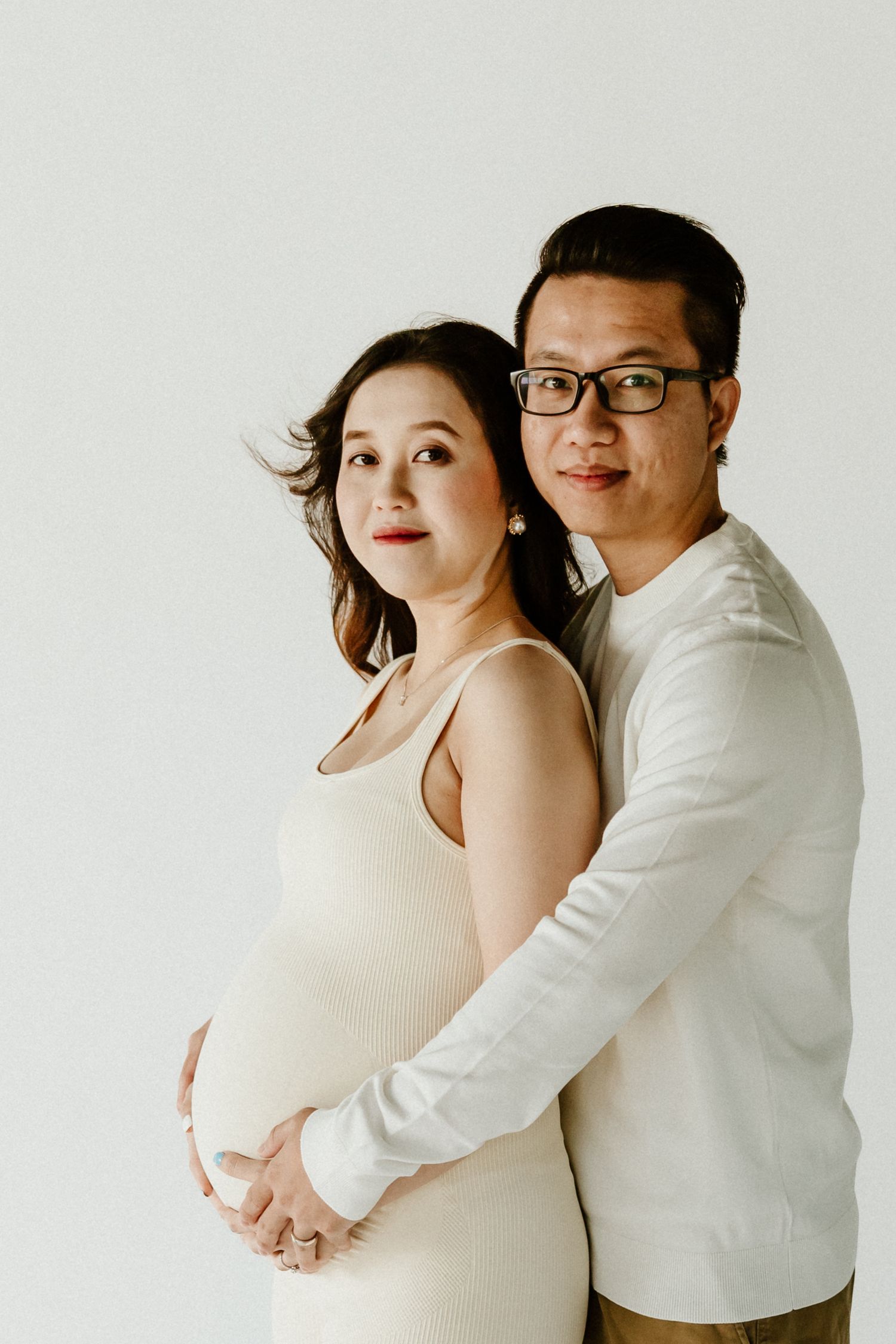 Simple studio maternity portrait with couple in white clothing against plain backdrop.