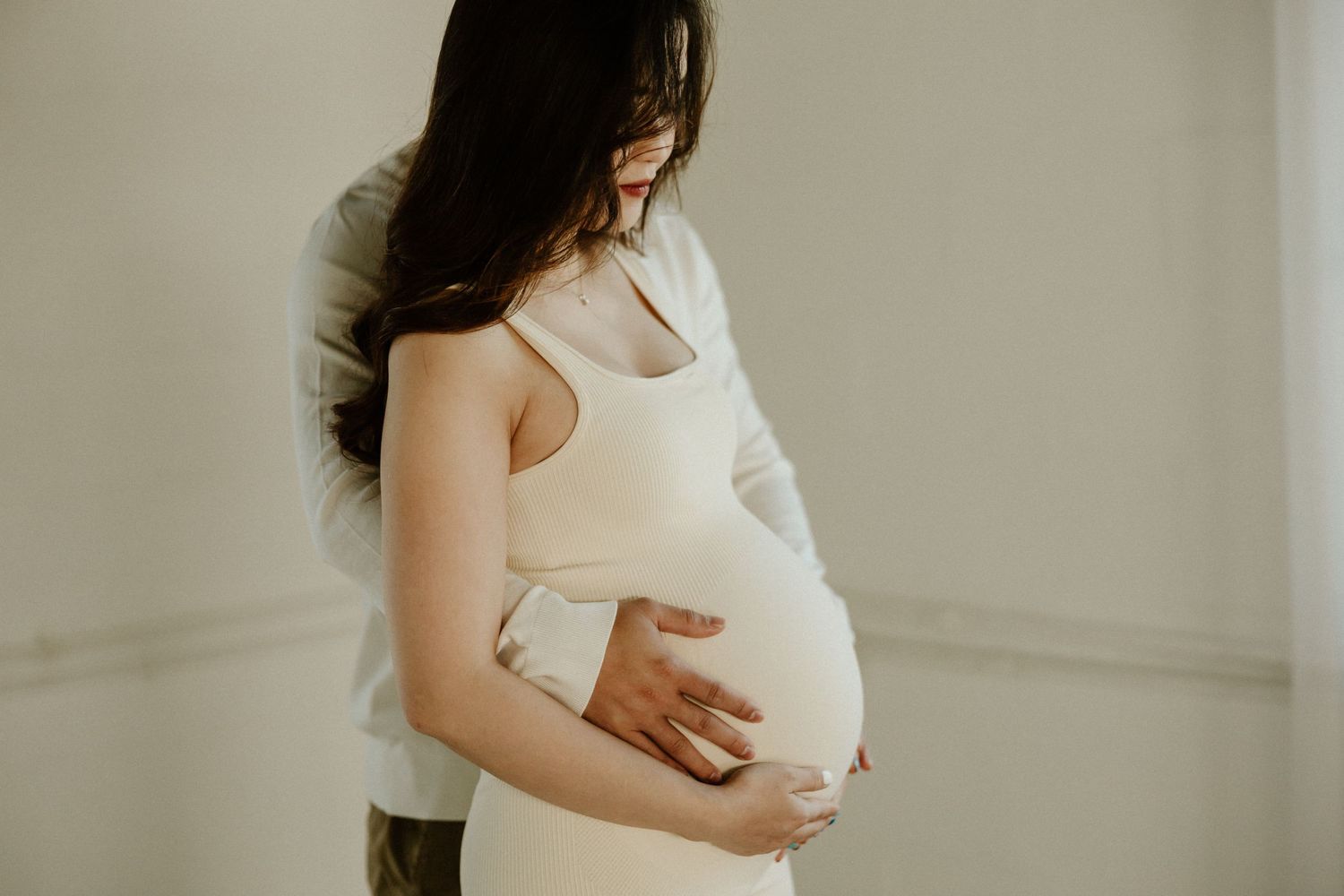 Side view of expectant couple embracing in front of white wall during intimate maternity photoshoot.