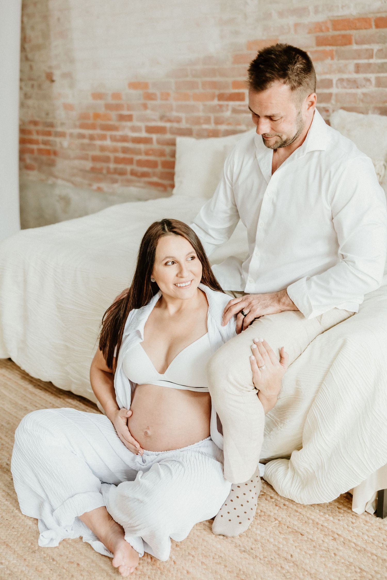 Intimate maternity photo session with couple in white outfits sitting on white couch against brick wall.