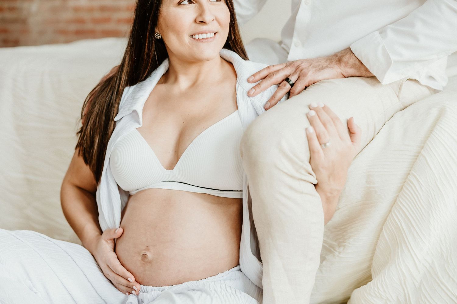 Expecting mother in white loungewear with visible baby bump sitting on couch during photoshoot.