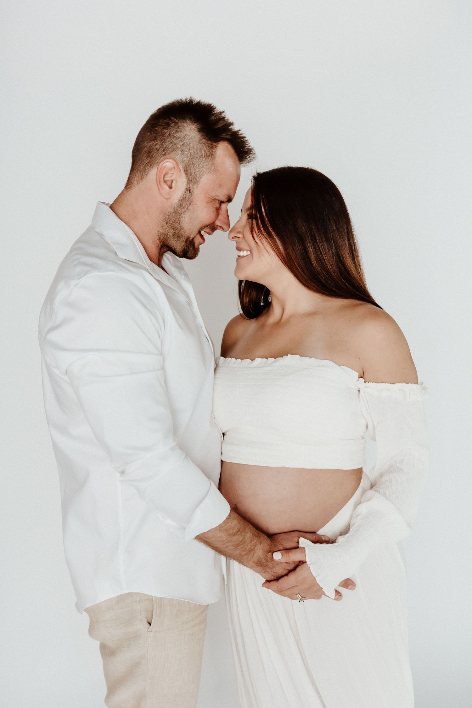 Romantic maternity moment with couple in white outfits touching foreheads against light background.