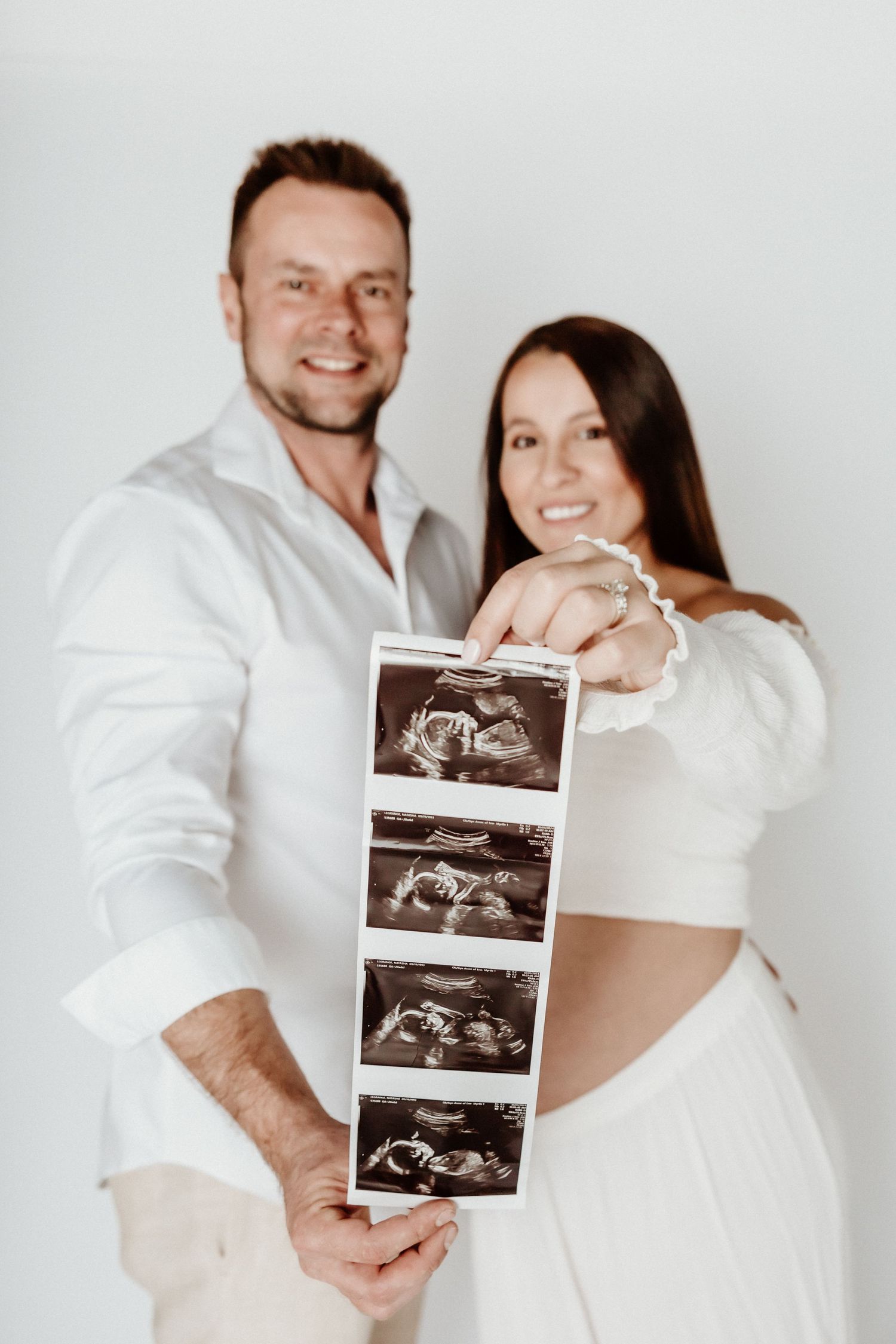 Expecting couple holding ultrasound images during pregnancy photoshoot against white background.
