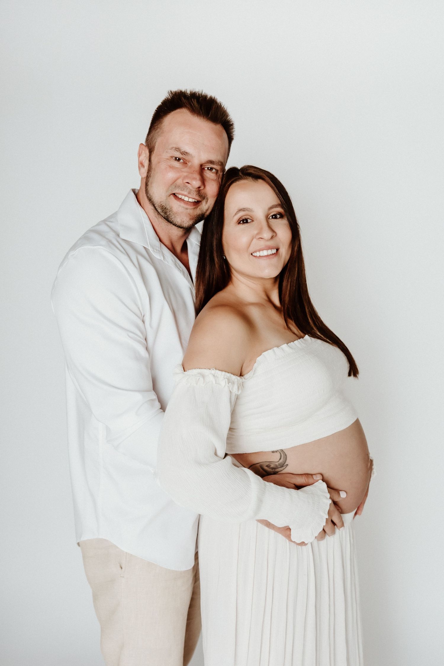 Maternity portrait of couple wearing white and beige against neutral background.