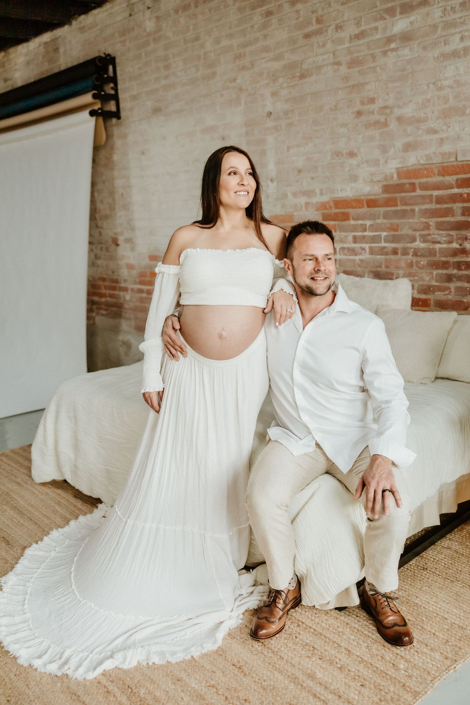 Maternity photo featuring couple in white attire posing on couch against brick wall.