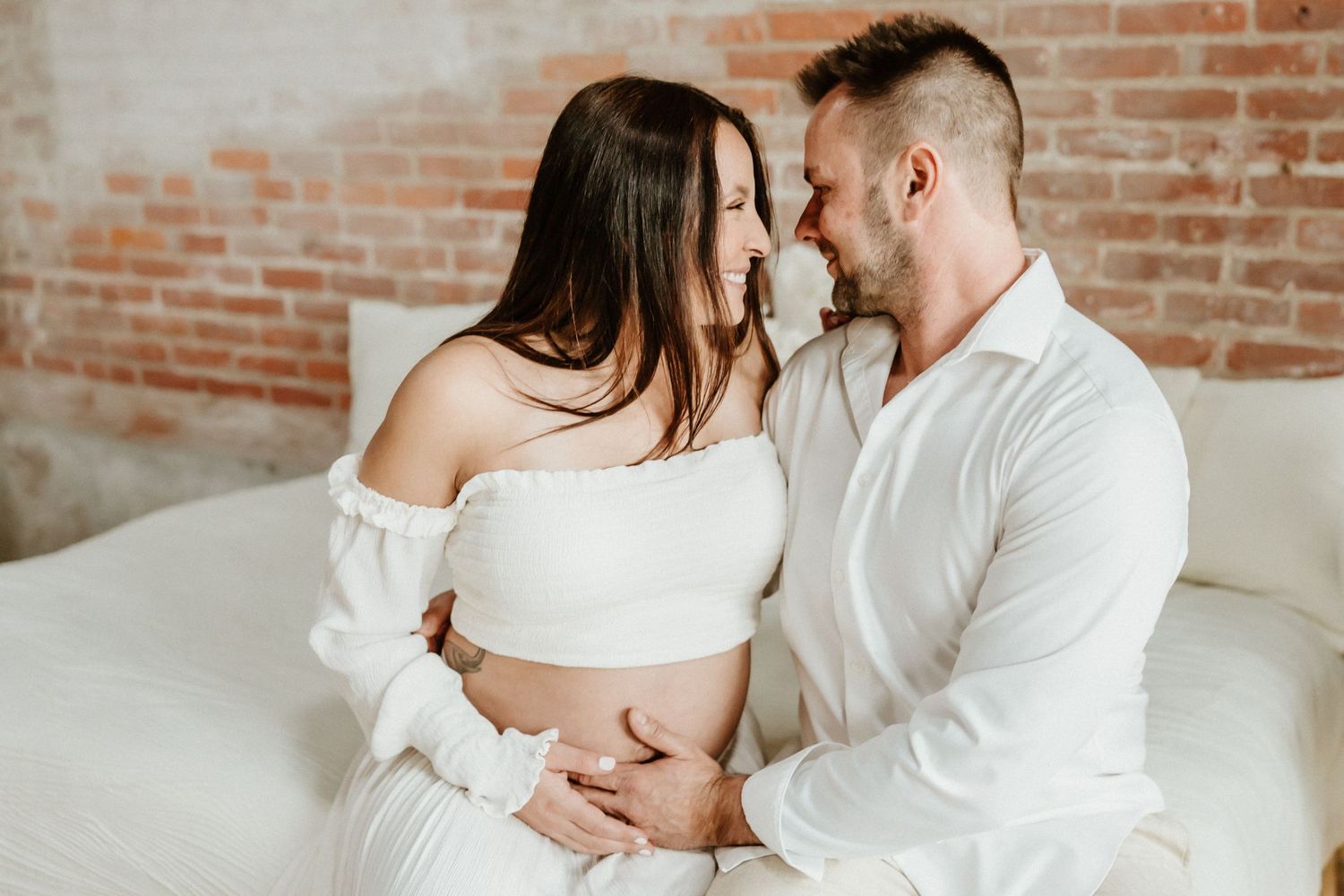 Intimate maternity moment with couple in white sharing tender moment on white couch.