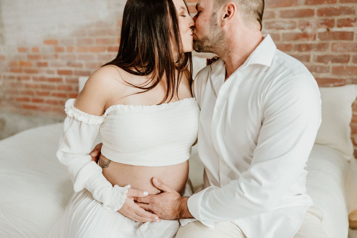 Tender pregnancy photo of couple in white embracing on couch during maternity session.