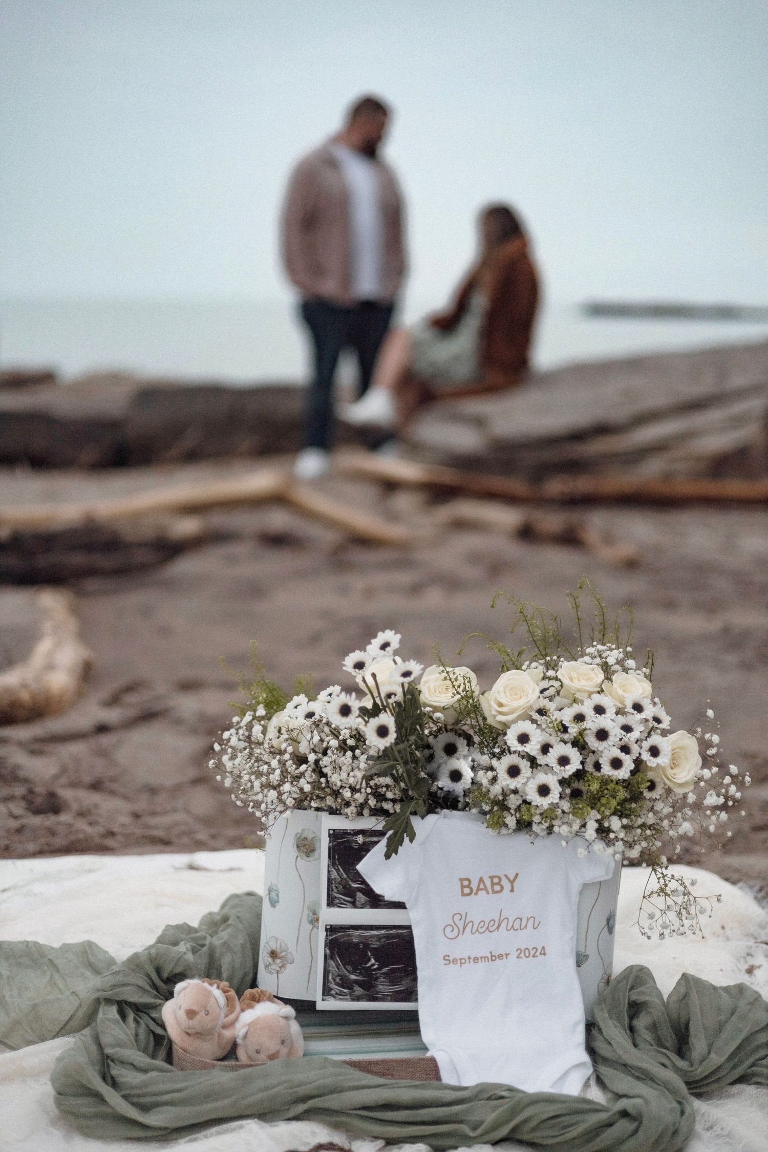 Baby announcement display with ultrasound photo and white flowers on a beach with couple silhouetted in background.