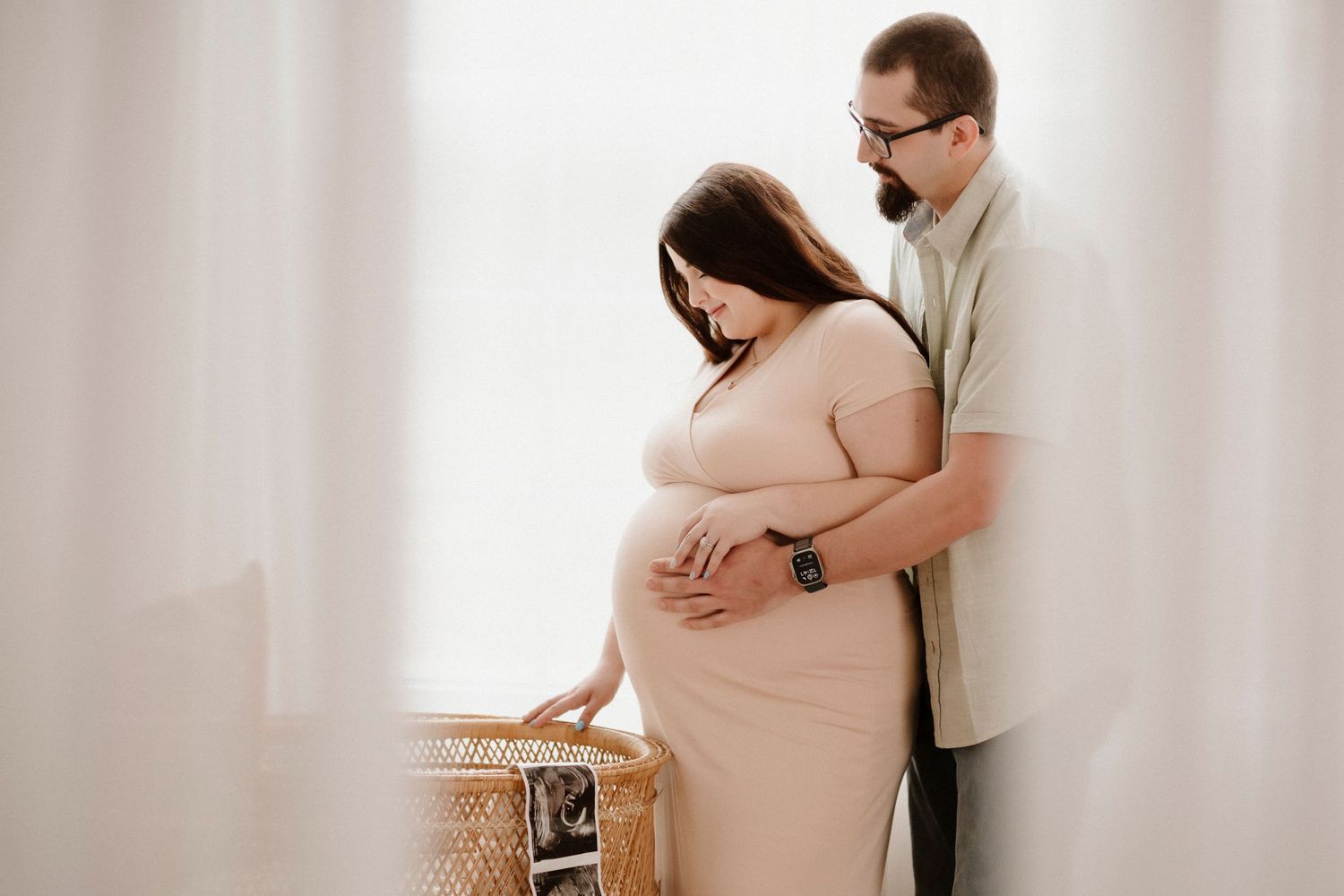 Intimate maternity photo of couple embracing near window with woven decor.