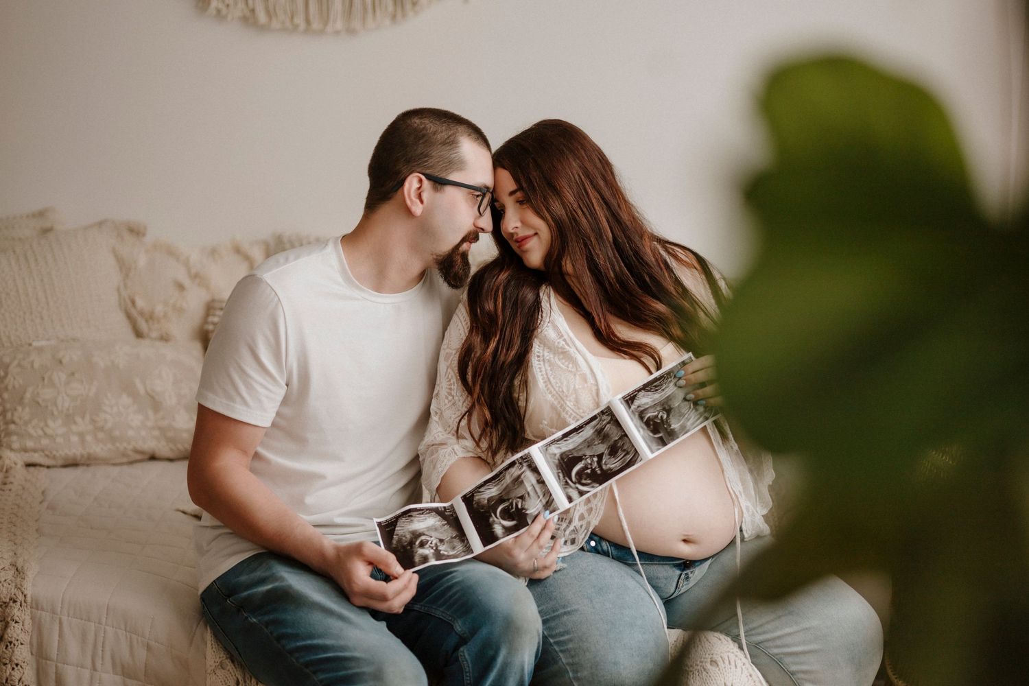 Couple shares tender moment while looking at strip of ultrasound photos on beige couch.