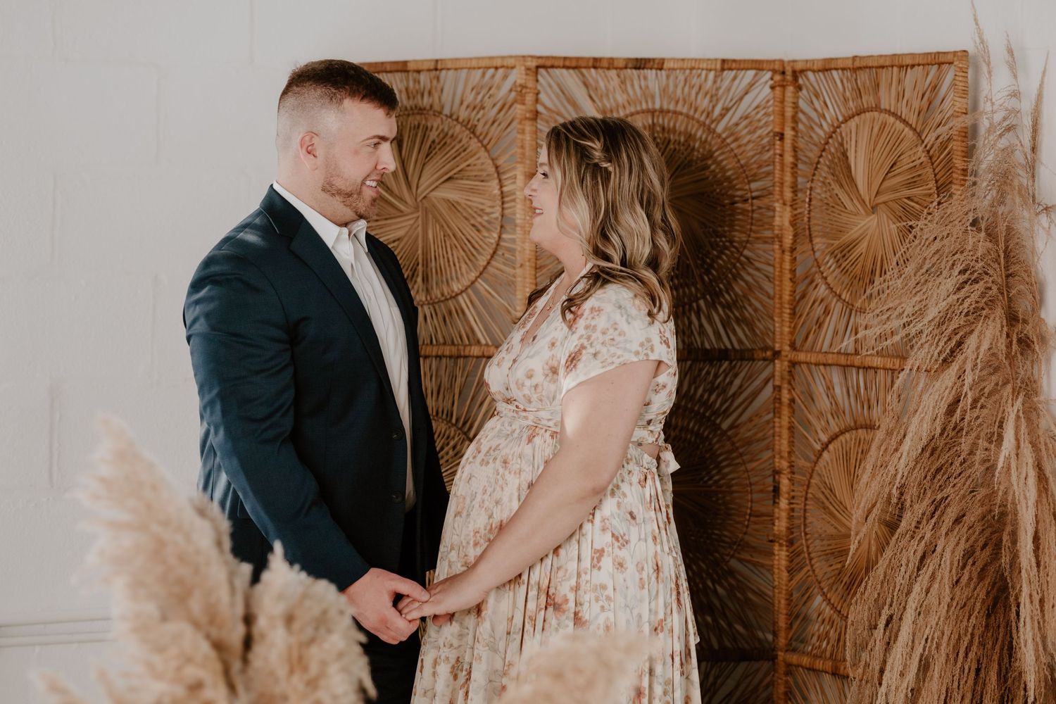 Couple shares tender moment against woven rattan backdrop during maternity shoot.