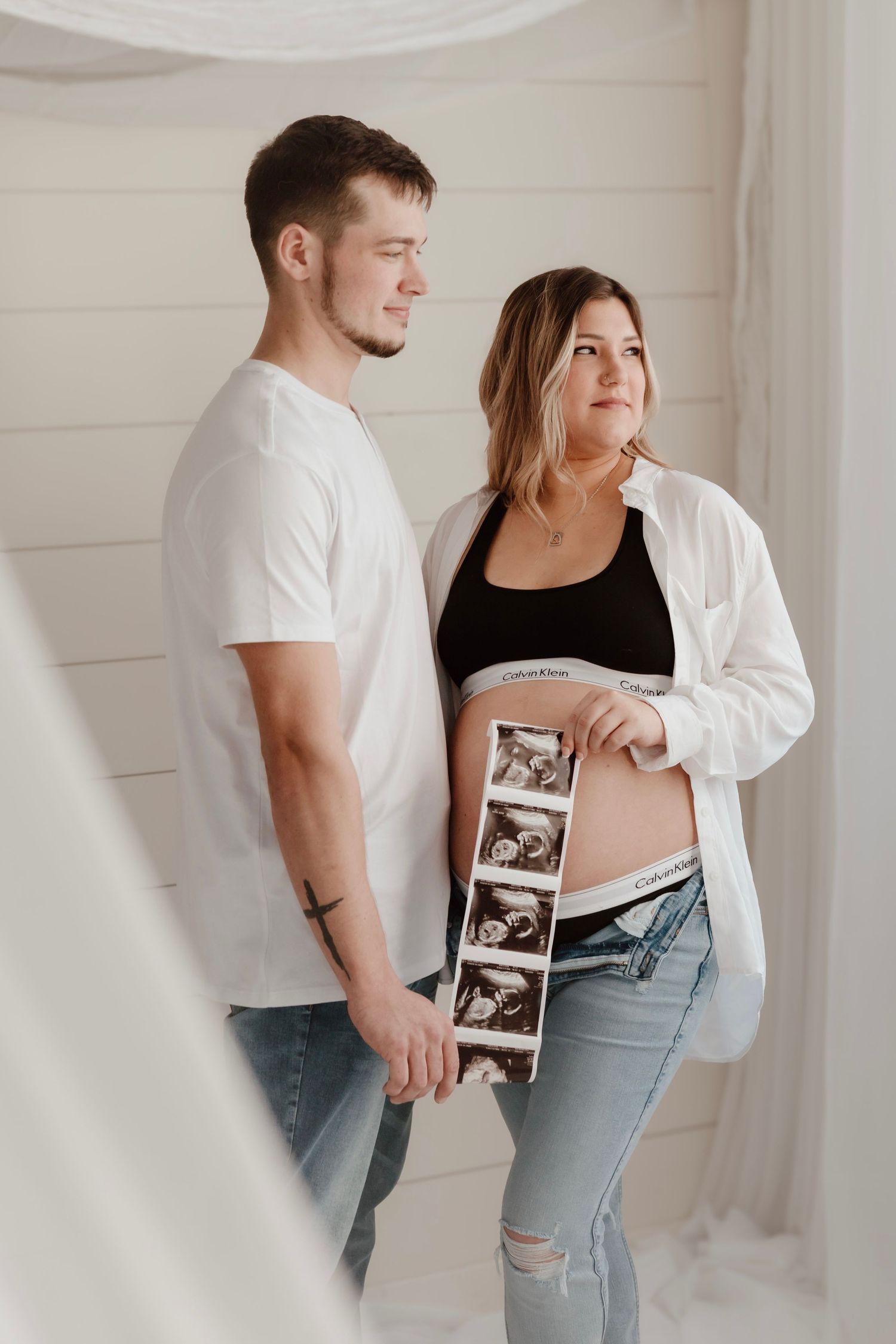 Expectant couple poses with ultrasound photos while wearing casual attire against white shiplap wall.