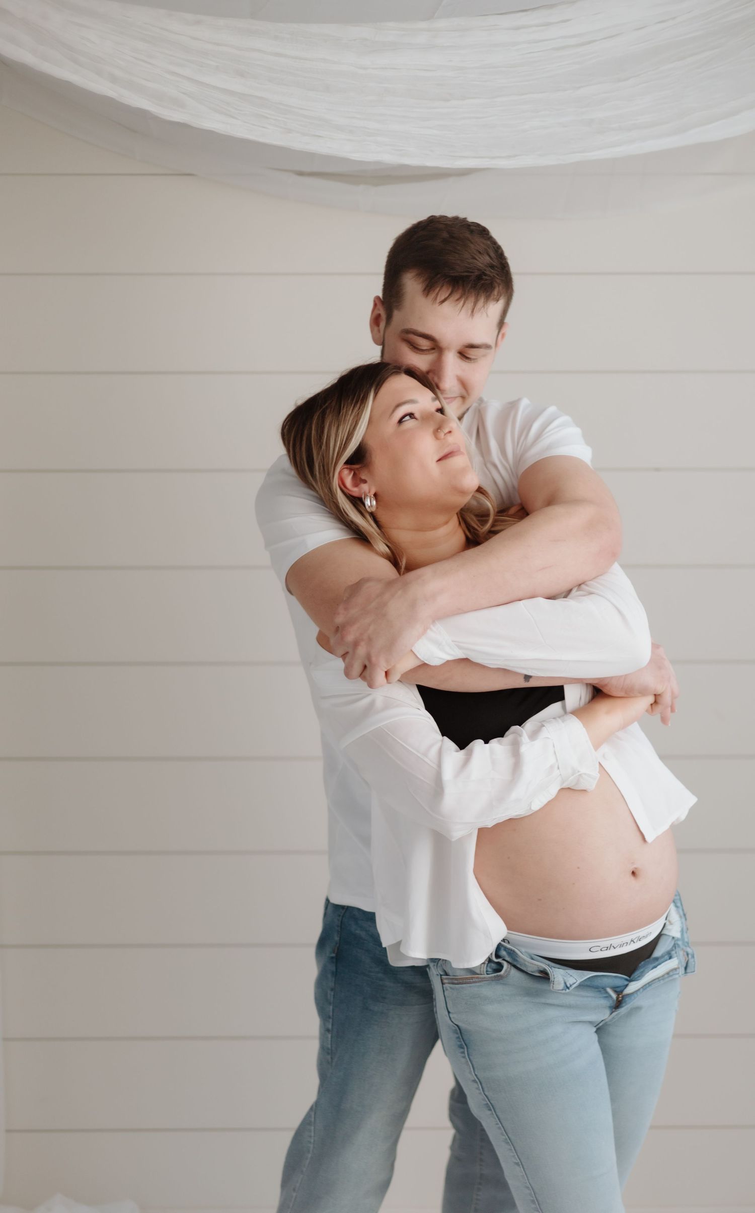 Intimate maternity photo of couple embracing against white wall.