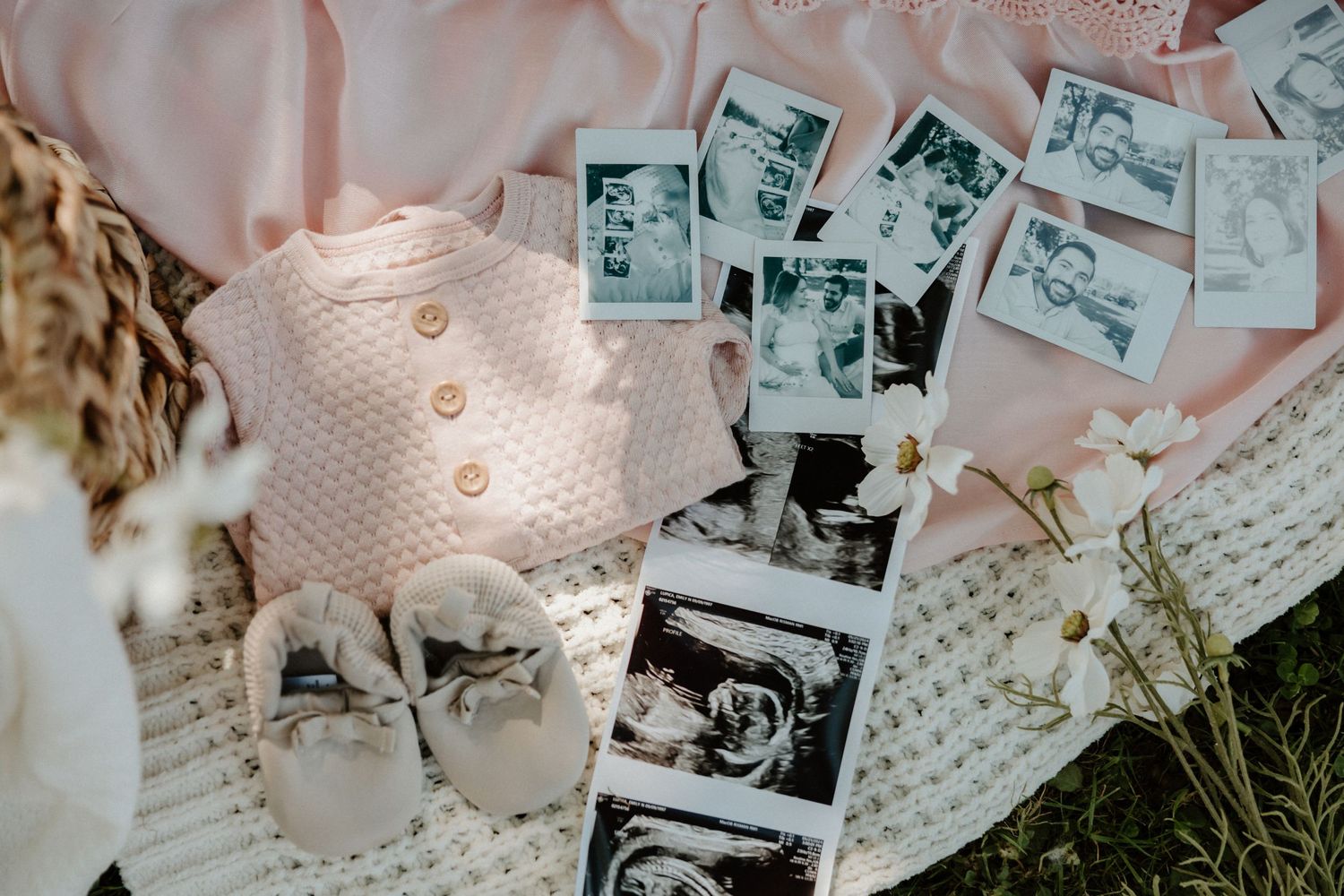 Vintage-style polaroid photos arranged on pink fabric with baby shoes and dried flowers.