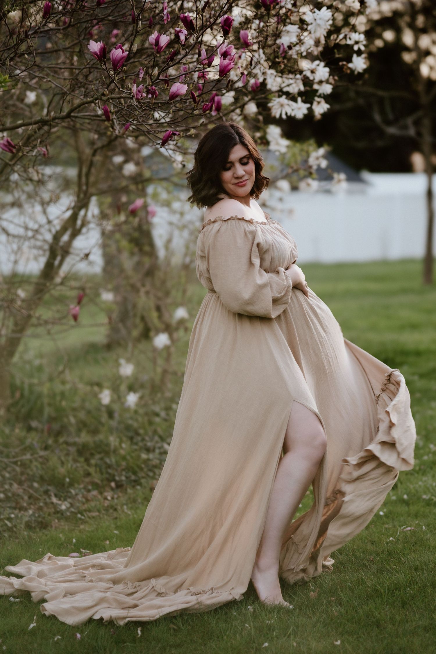 A flowing beige maternity dress twirls beneath blooming magnolia trees in spring.