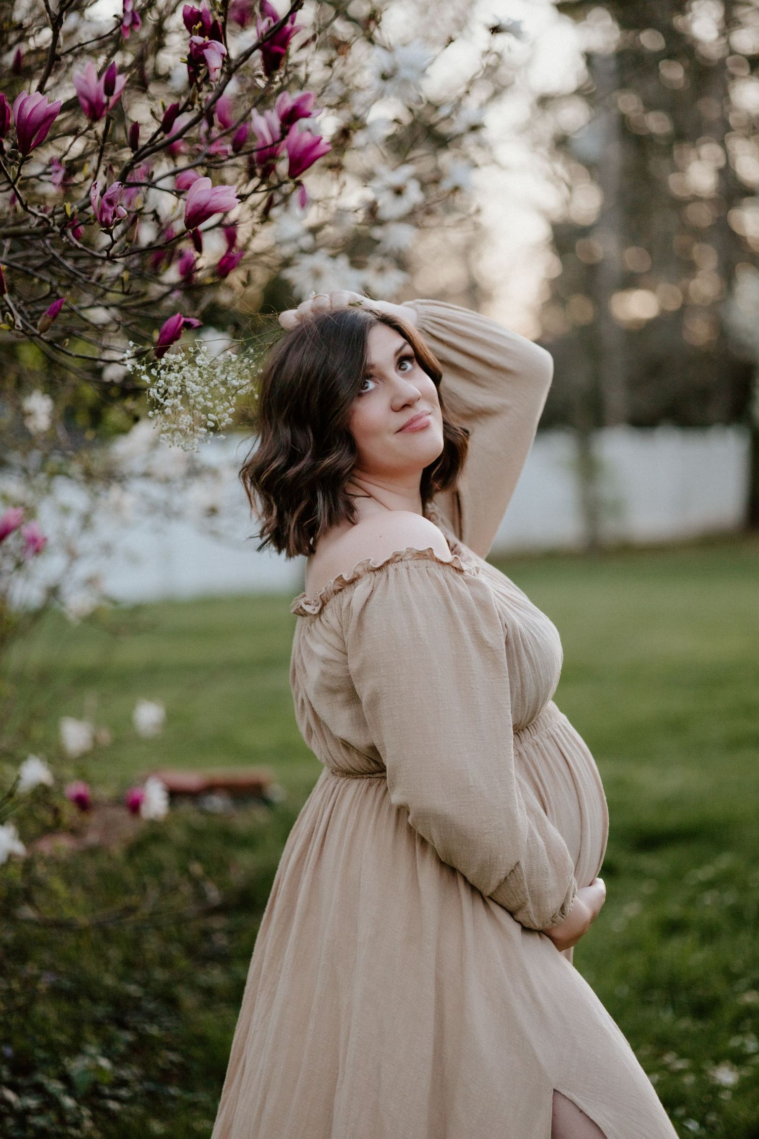 Expecting mother in beige dress poses elegantly under flowering magnolia branches.