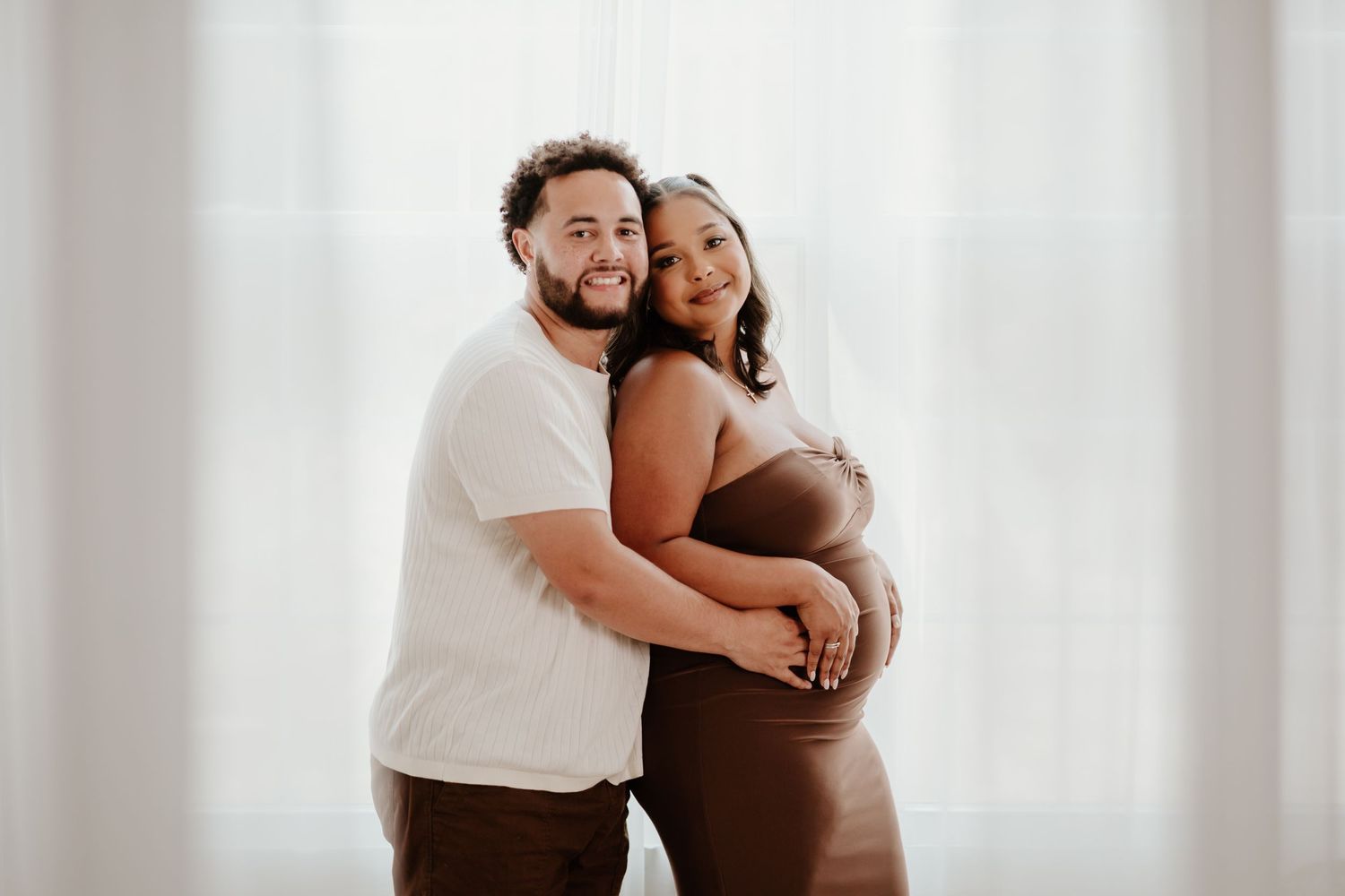 A couple embraces during a maternity photoshoot against a white backdrop.