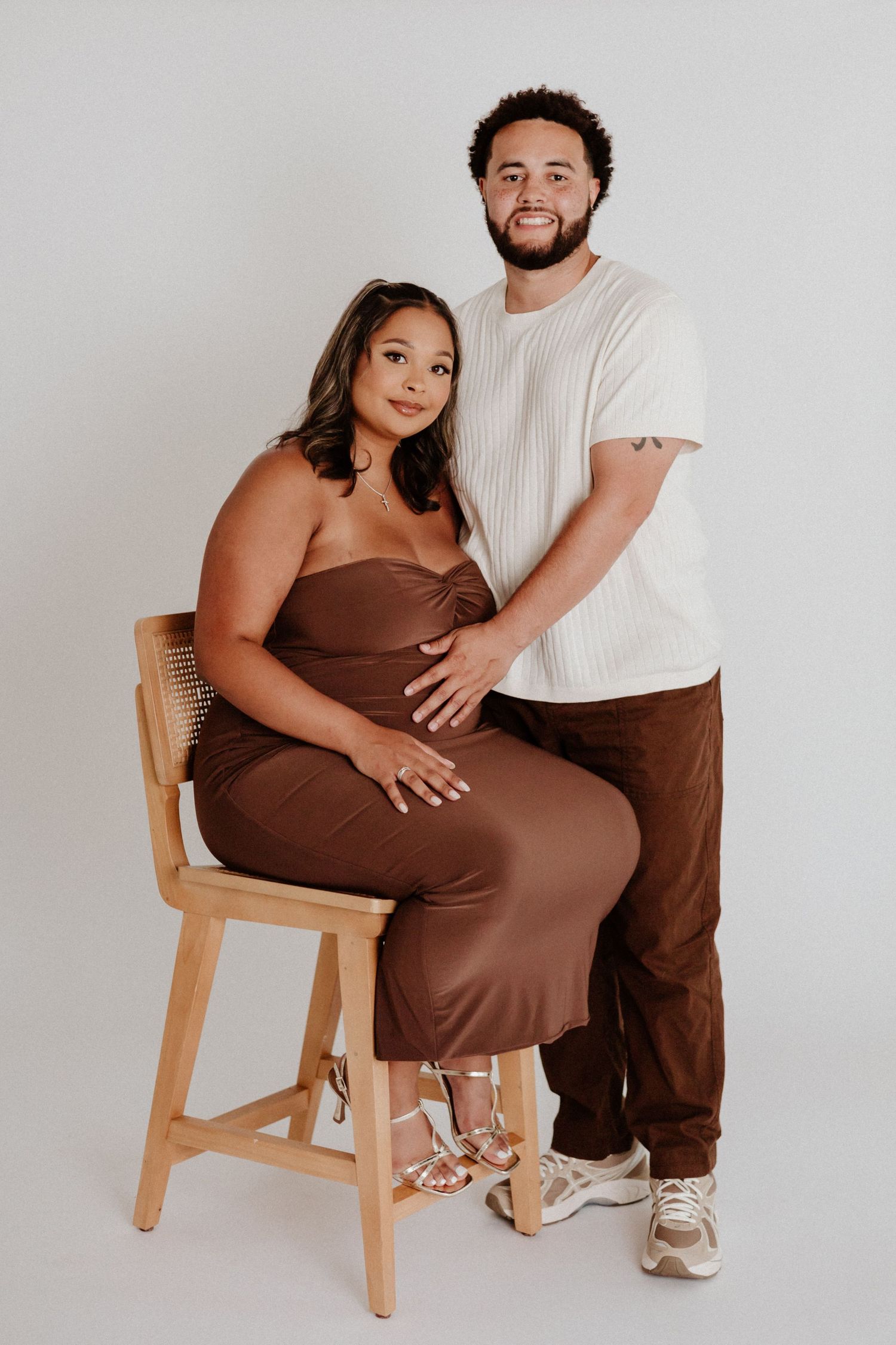 Maternity portrait of couple by white wall with woman in brown dress seated on wooden chair.