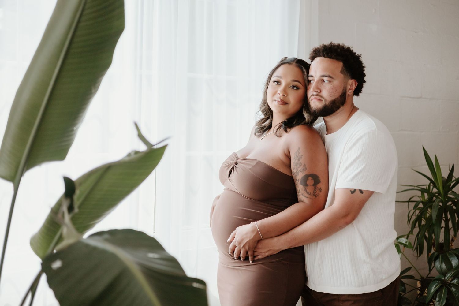 Maternity photo with tropical plants featuring couple in casual attire by bright window.