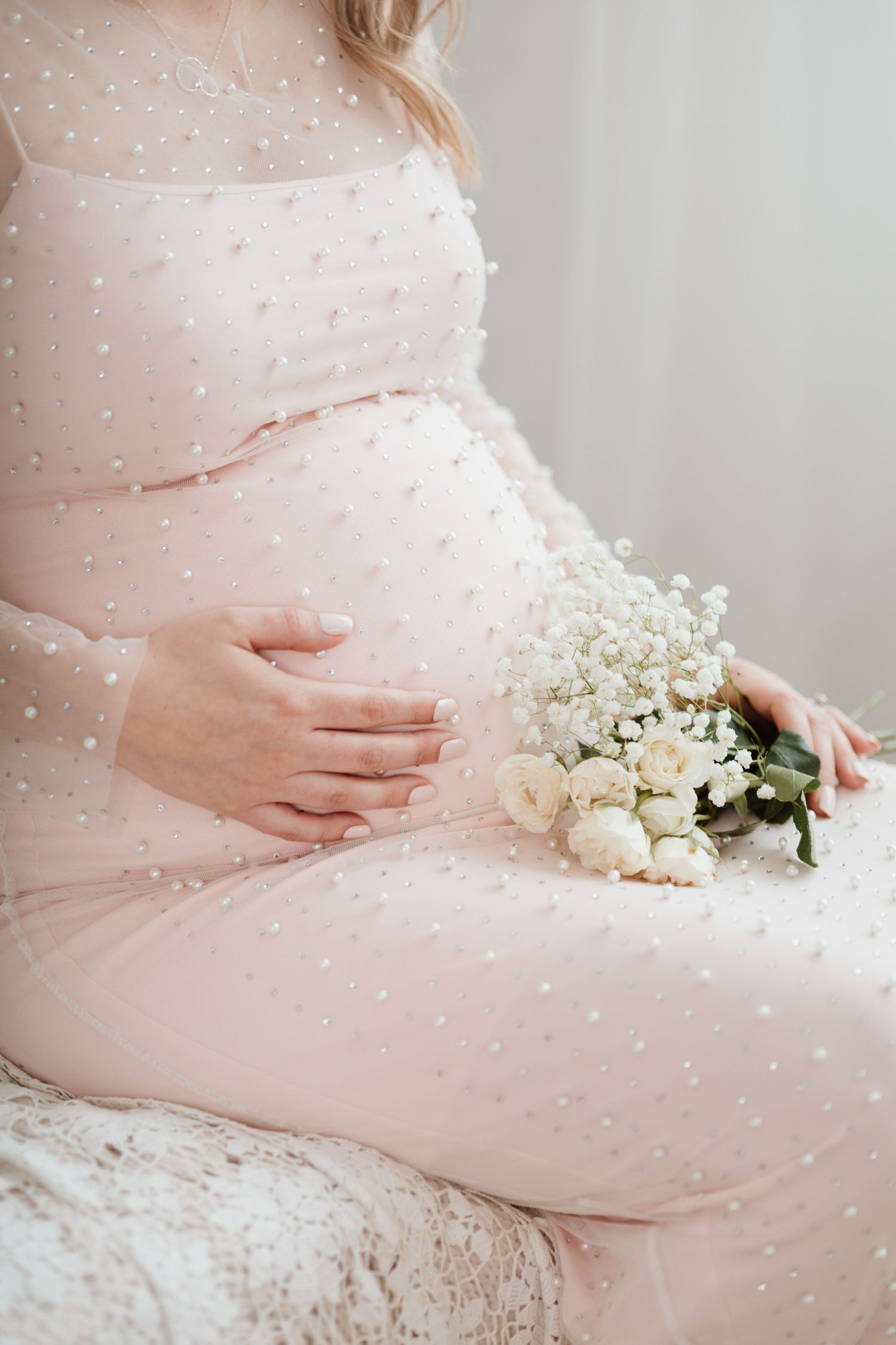 A pregnant woman in a pearl-dotted pink dress holding white flowers against a light background.
