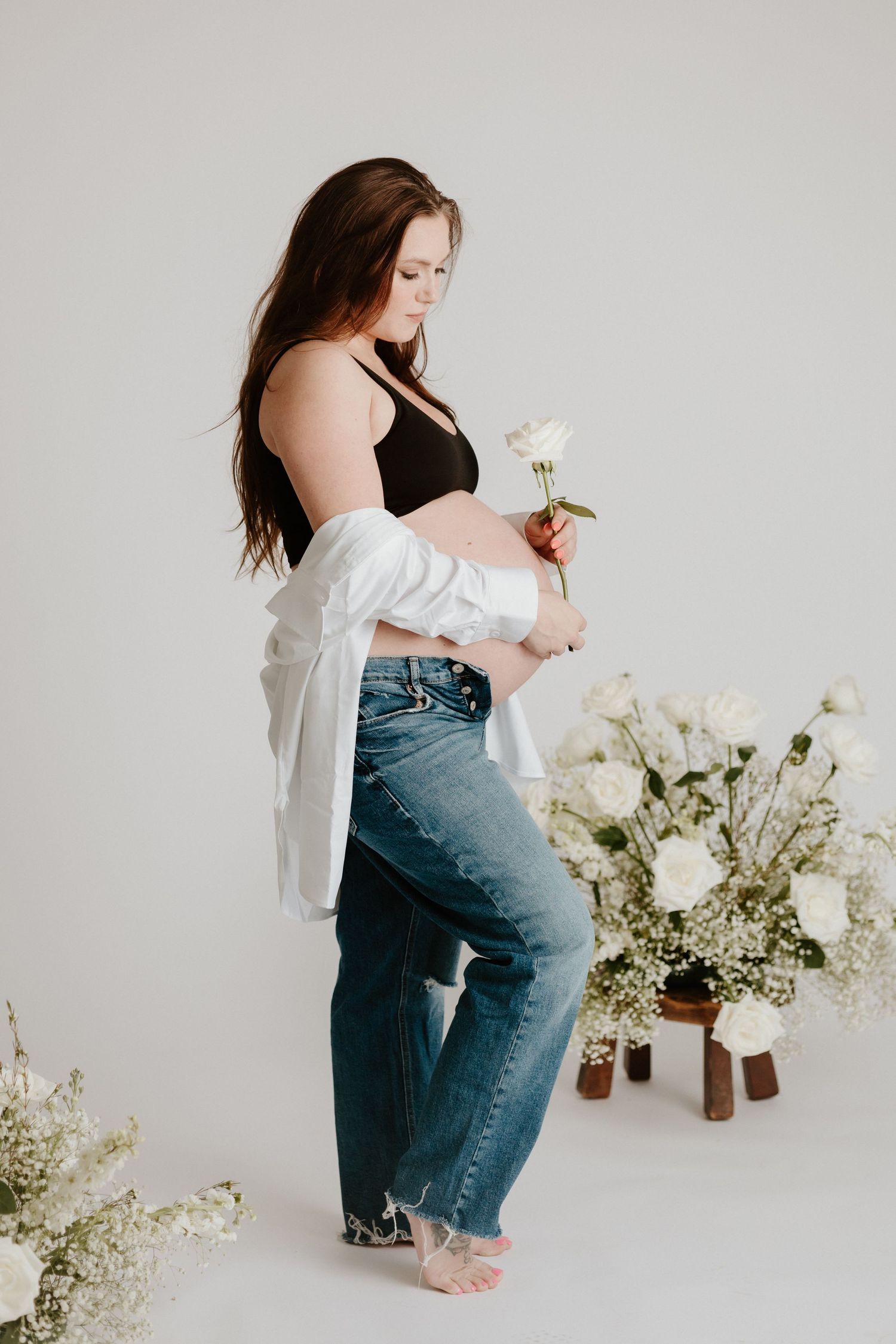 Artistic maternity photo with a woman in distressed jeans and white cardigan posed near white floral arrangements.