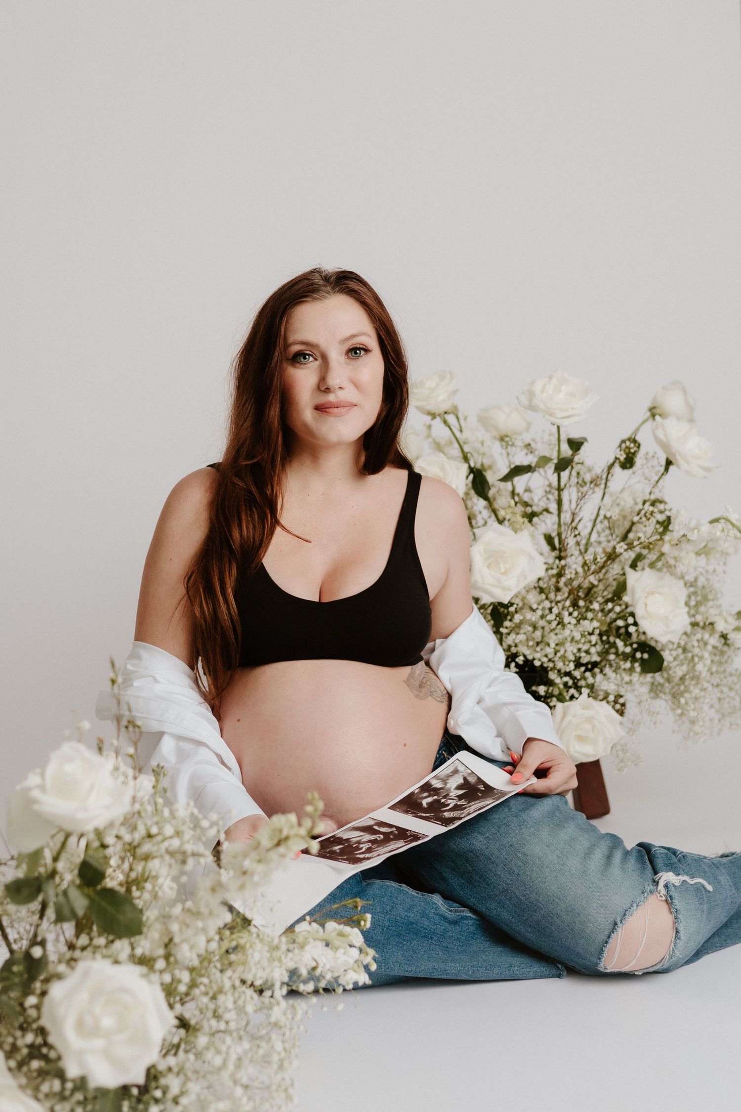 Maternity photo with white flowers showing an expectant mother holding ultrasound photos in casual attire.
