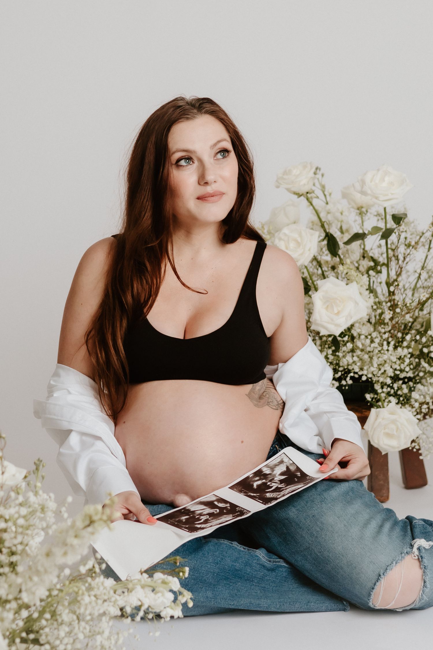 Studio maternity portrait with white flowers showing an expectant mother holding ultrasound photos.