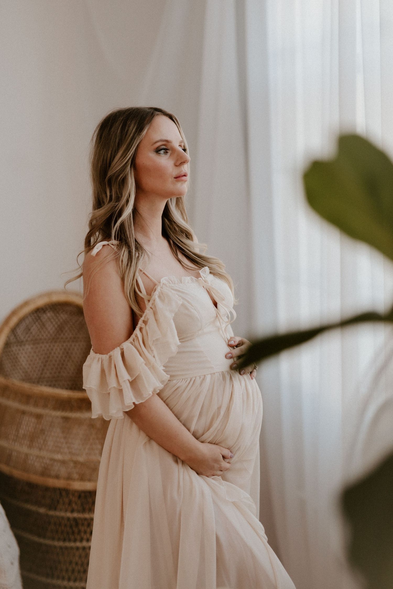Expectant mother in flowing cream dress poses elegantly against sheer curtains and woven peacock chair.
