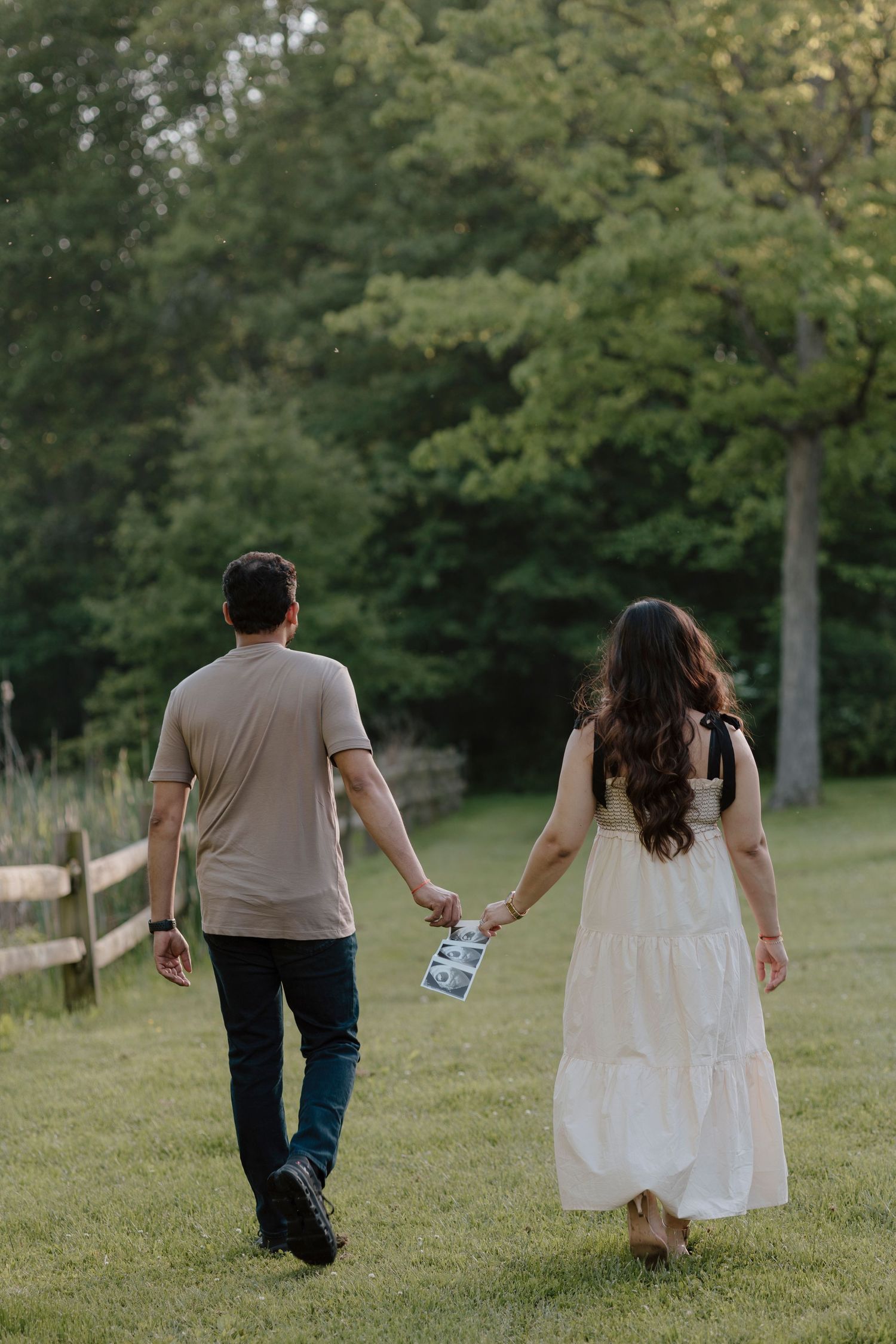 A couple walking hand in hand on a grassy path while holding ultrasound photos.