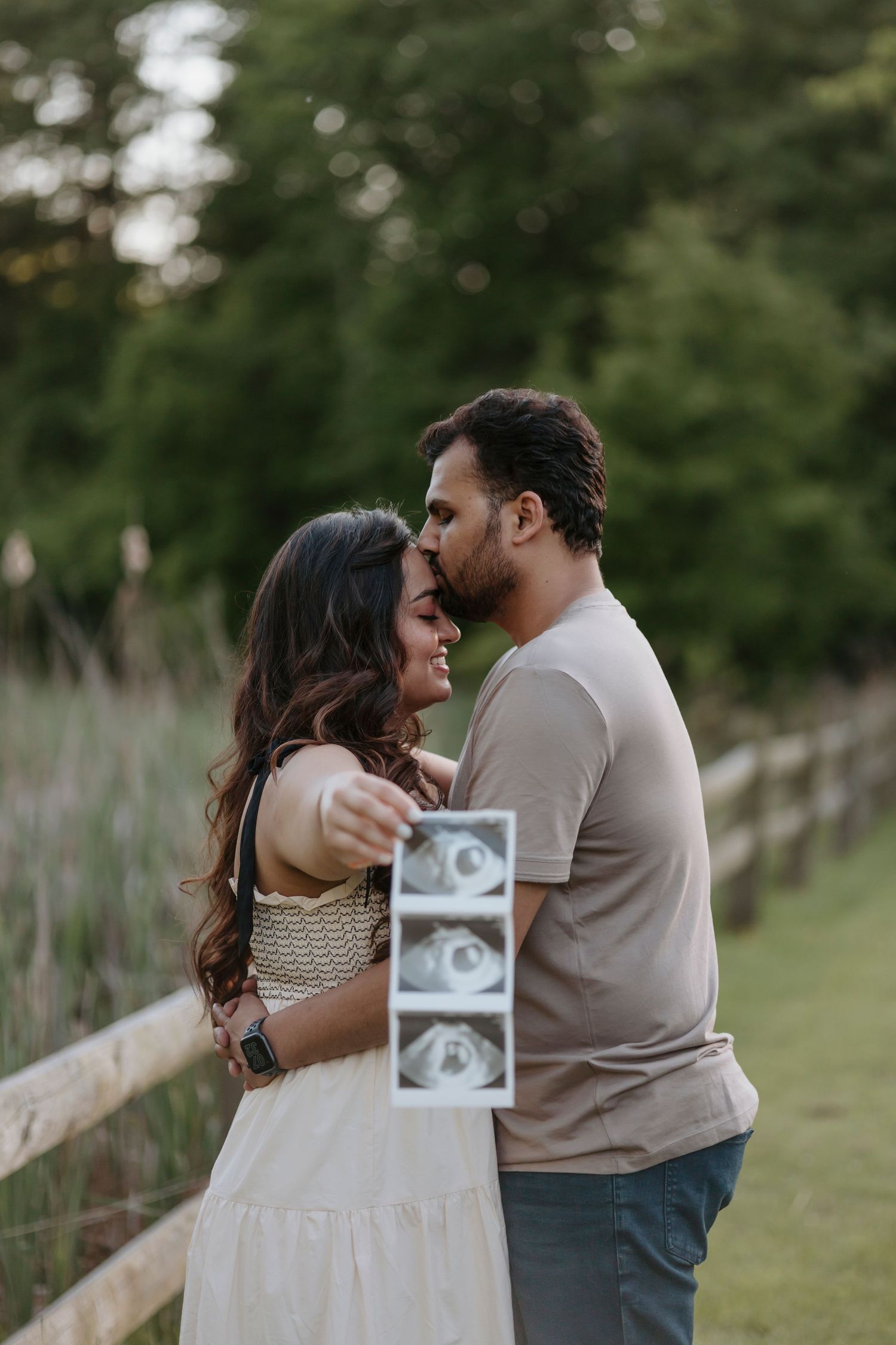 A couple sharing an intimate embrace while holding ultrasound photos in an outdoor setting.