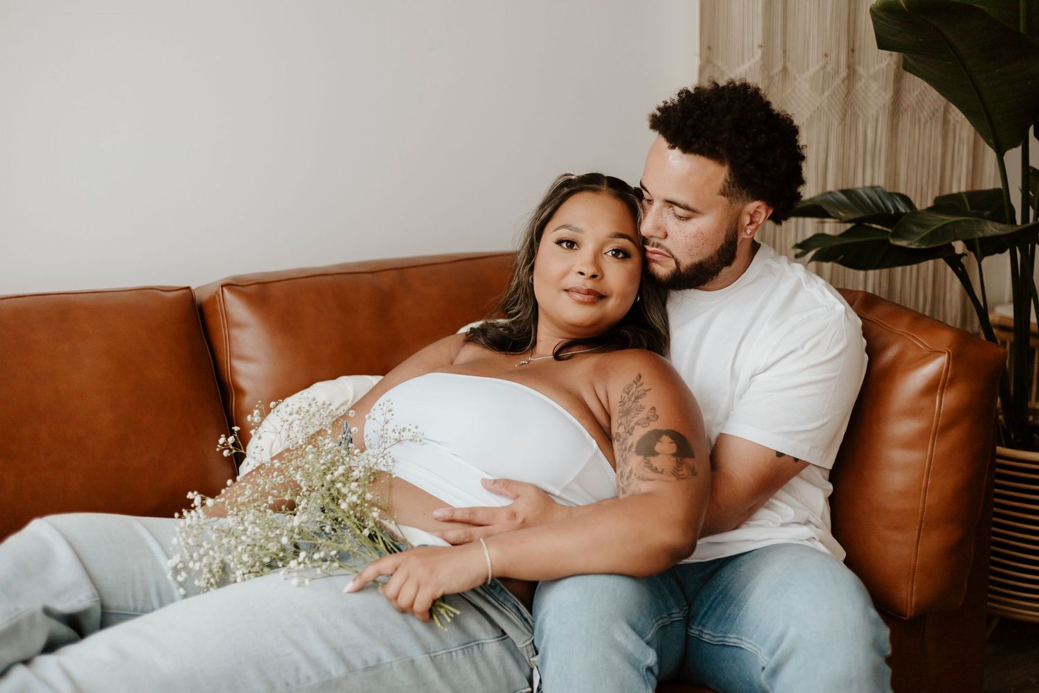 A maternity photo of a couple cuddling on a tan leather sofa with flowers.