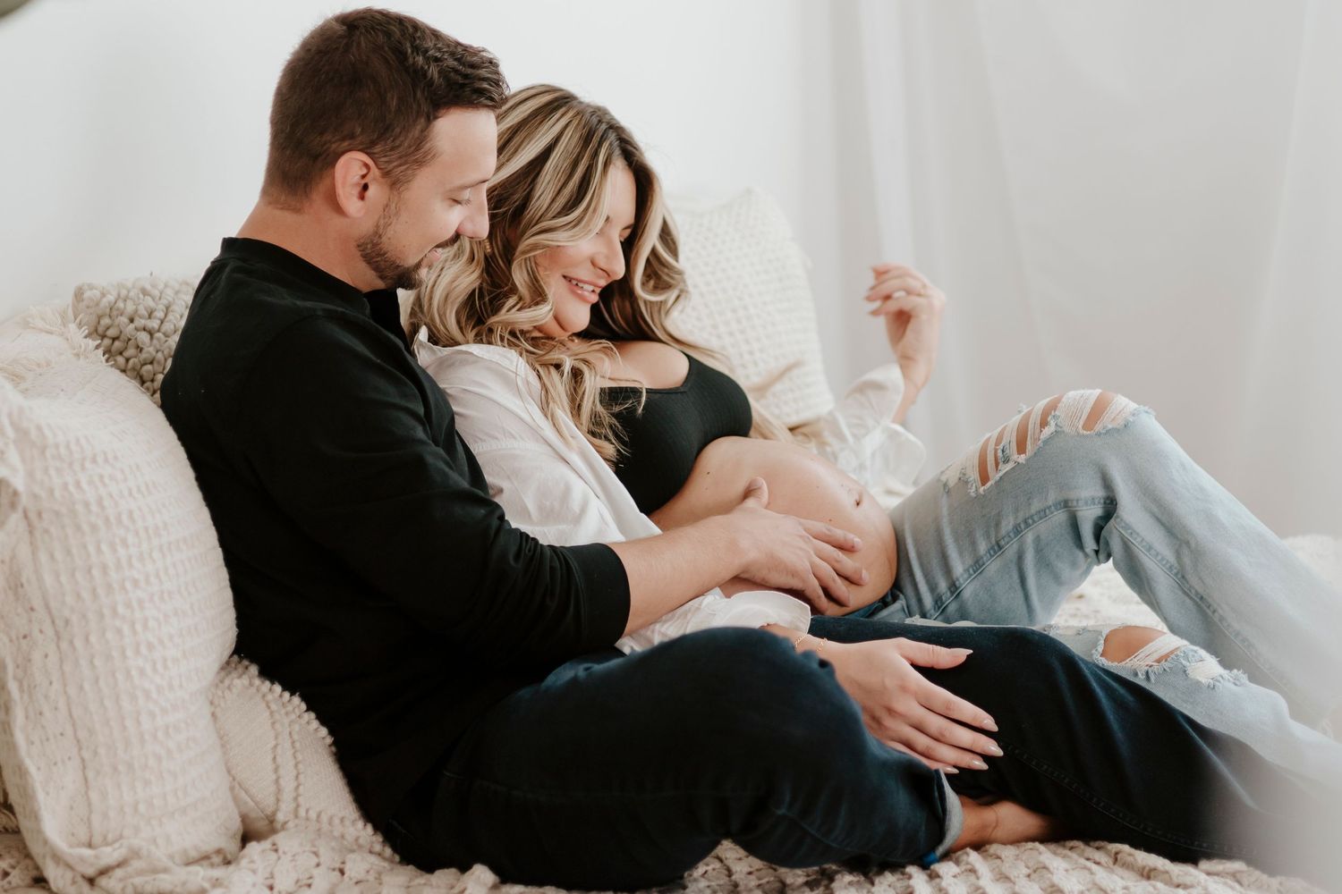 Expecting couple shares relaxed moment together on white couch in casual attire.