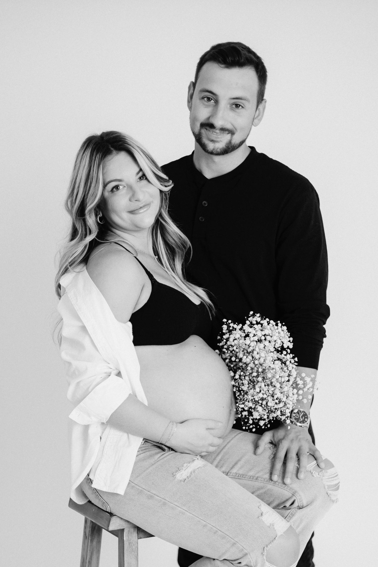 Maternity portrait with couple in contrasting outfits holding baby's breath flowers against white backdrop.