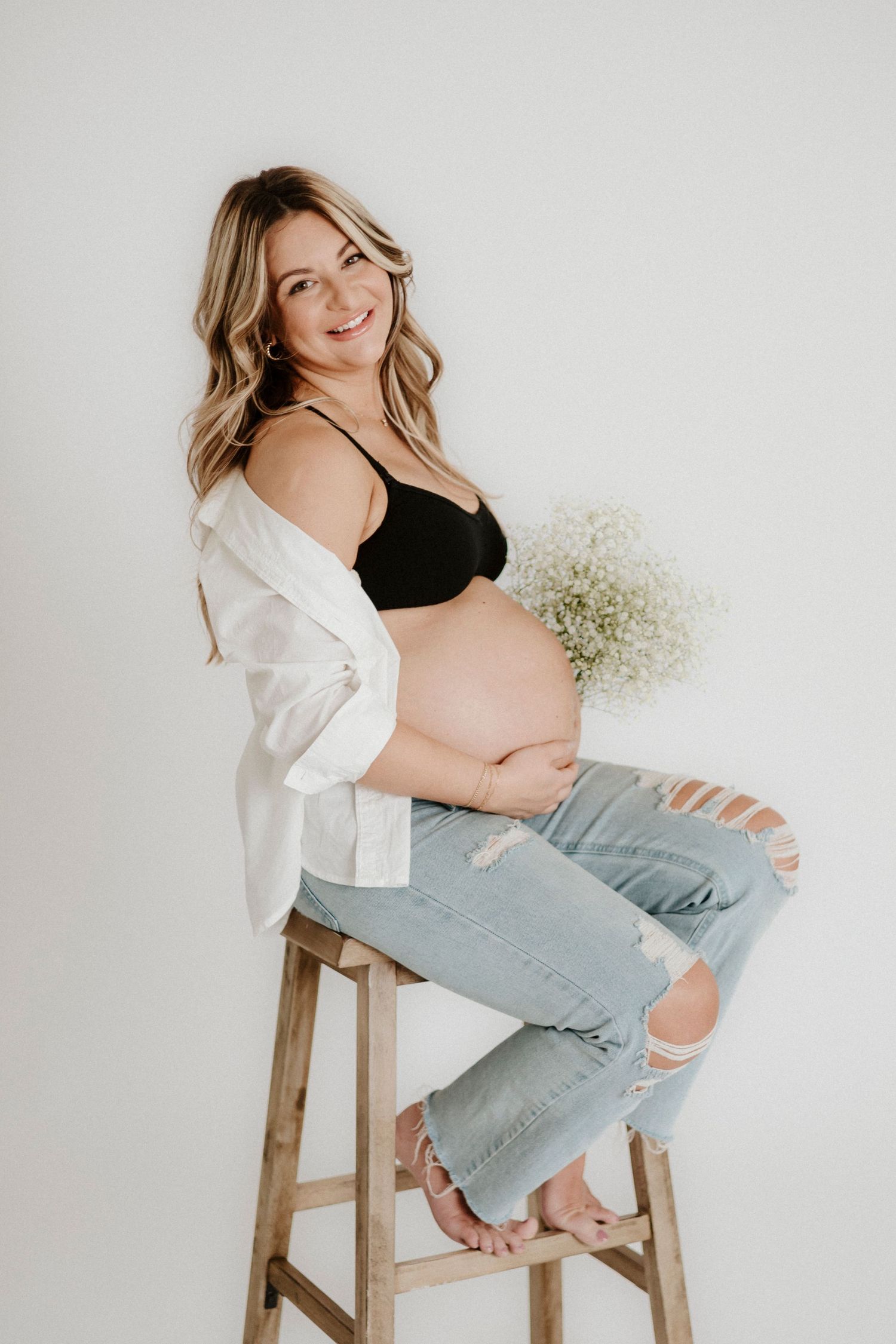 Expectant mother in ripped jeans and black top sits on wooden stool holding baby's breath flowers against white backdrop.