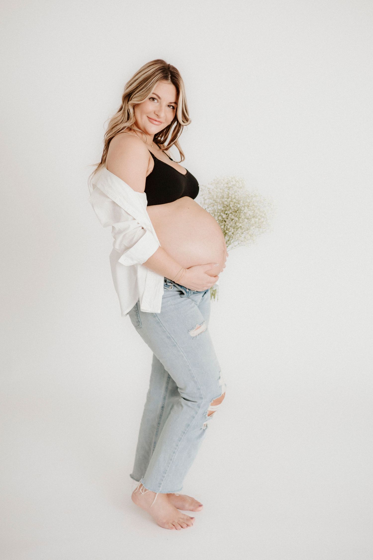Pregnant woman in casual attire poses standing against white background with delicate baby's breath flowers.
