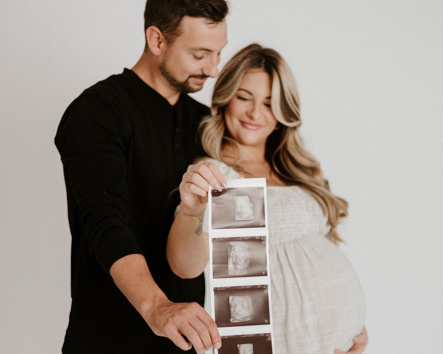 Expecting couple shares intimate moment while holding ultrasound photos against white background.