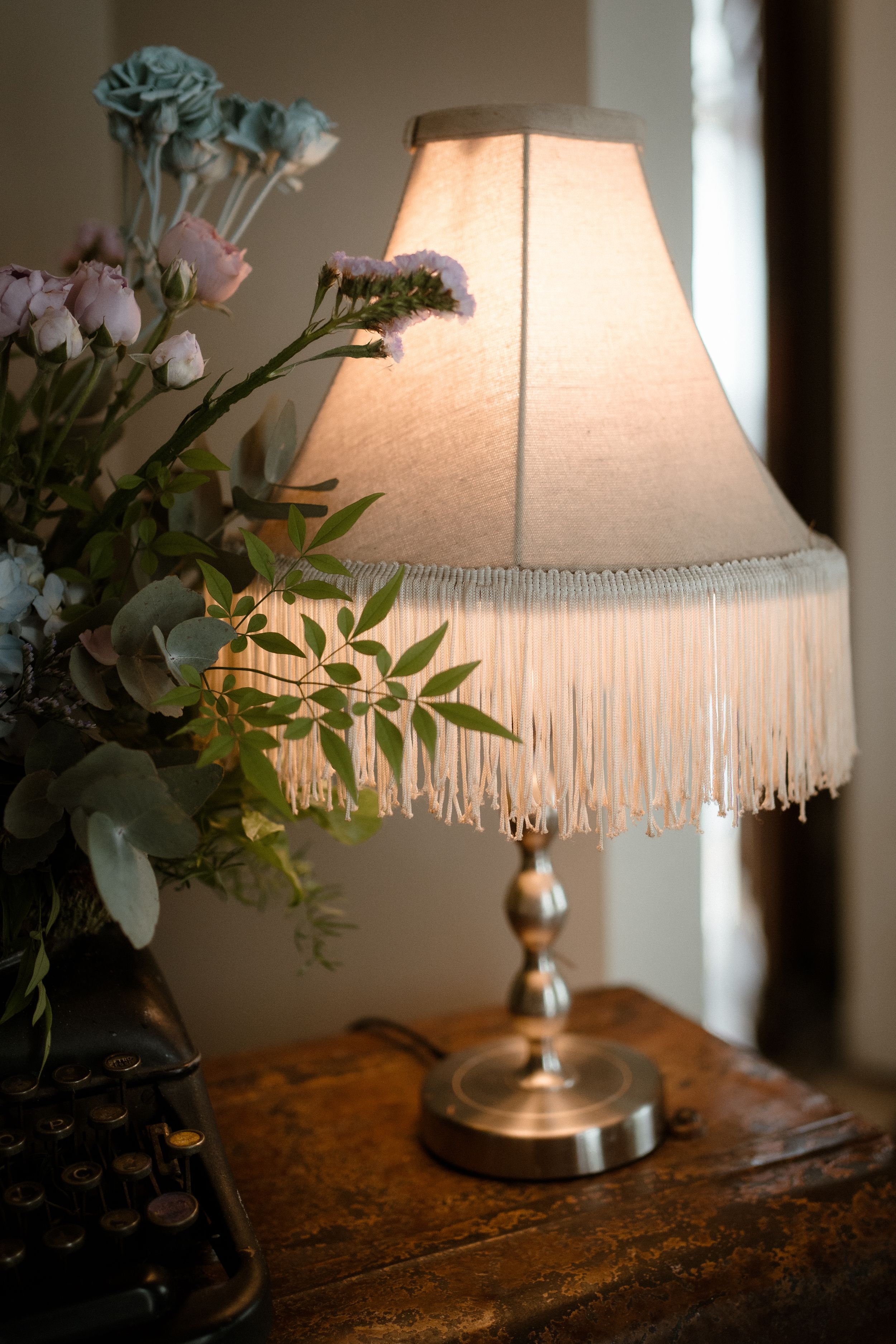 Vintage table lamp with fringed shade surrounded by delicate floral arrangements.