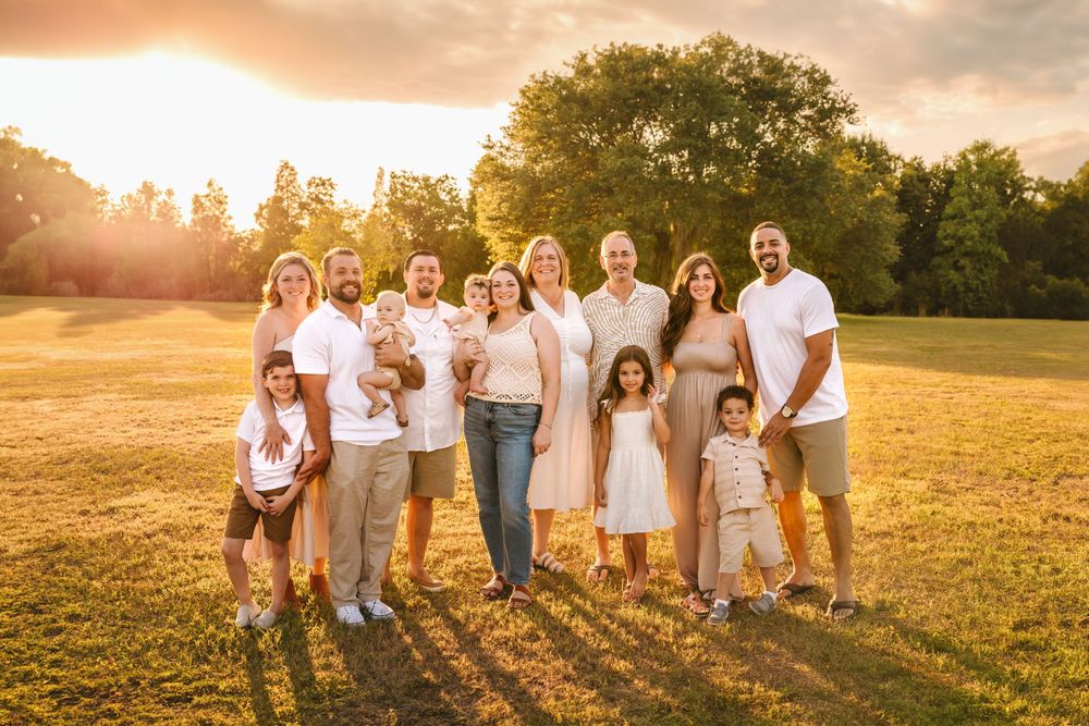Large family portrait at golden hour in a green field, smiling across three generations in coordinated neutral outfits.