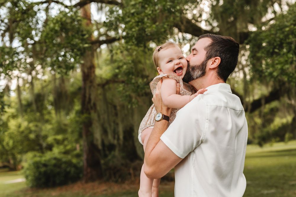 Joyful father lifting laughing baby girl high in a lush green park under a canopy of trees.