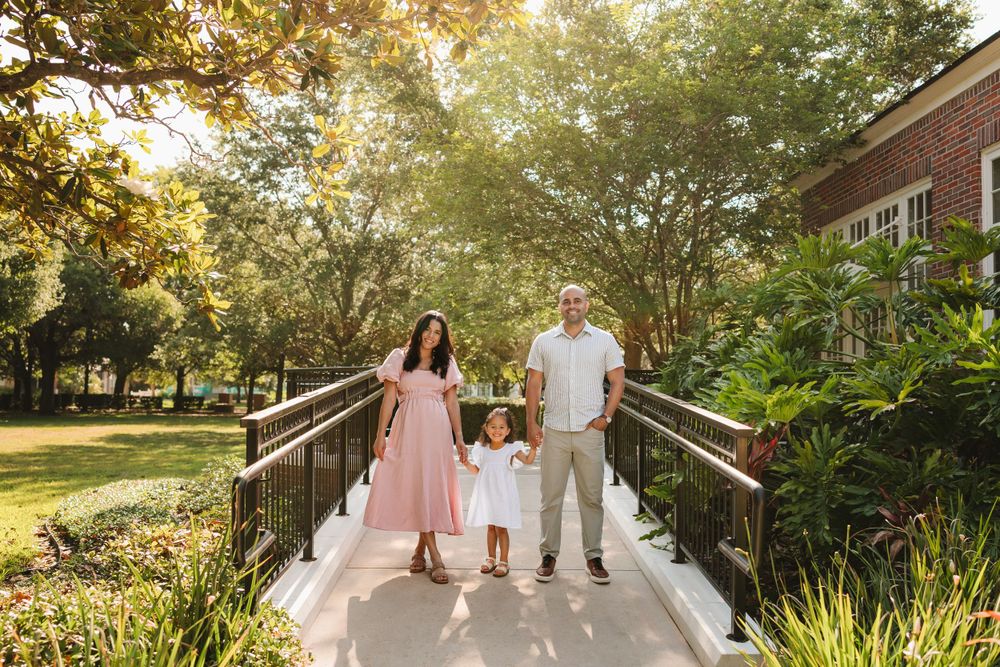Family of three holding hands on a sunlit garden bridge surrounded by lush greenery.