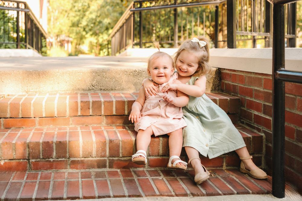 Two adorable little sisters hugging on brick steps, wearing pink and mint dresses in warm sunlight.