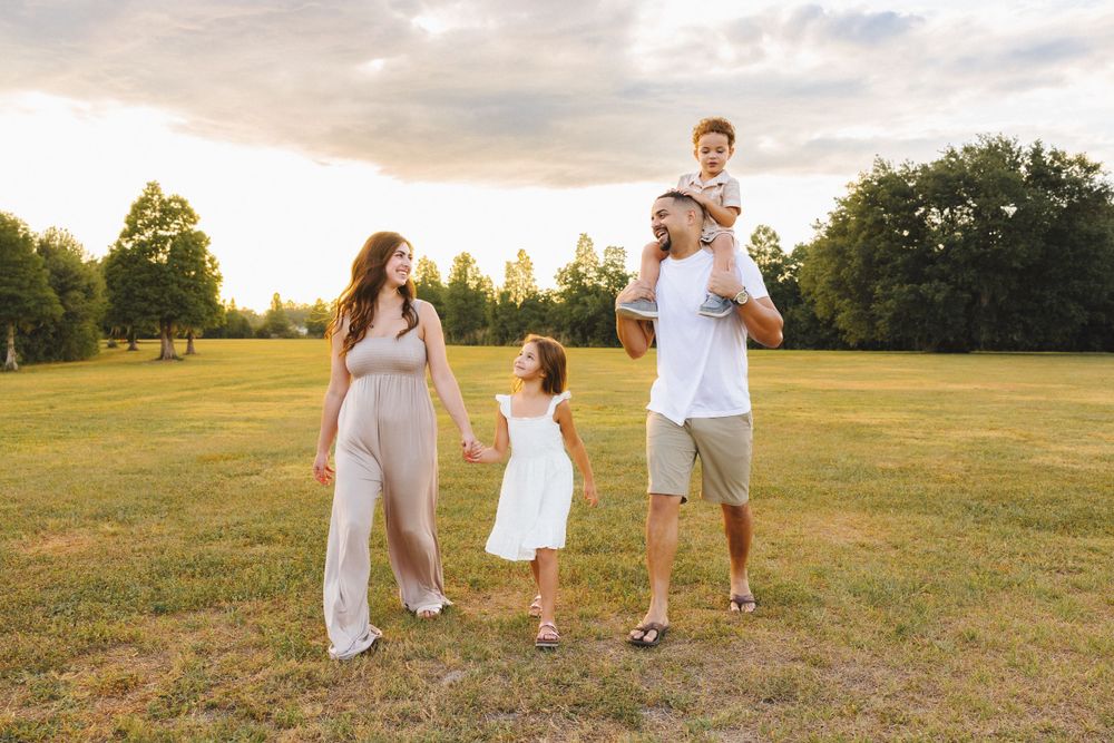 Happy family of four walking in a sunlit green field at golden hour, parents with two young children enjoying outdoors.