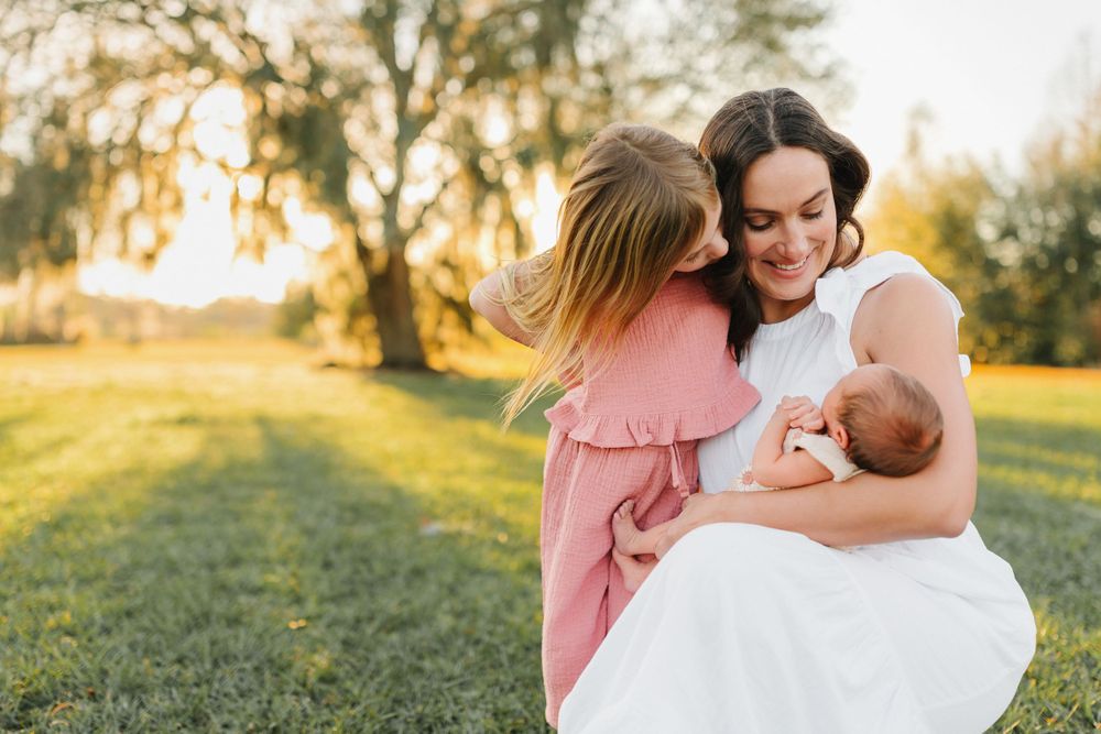 Smiling mother in white dress holds newborn baby while young girl in pink dress gives her a sweet kiss outdoors.