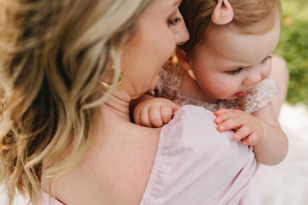 Blonde mother kissing her baby girl on the cheek, with infant clutching her shoulder gently.