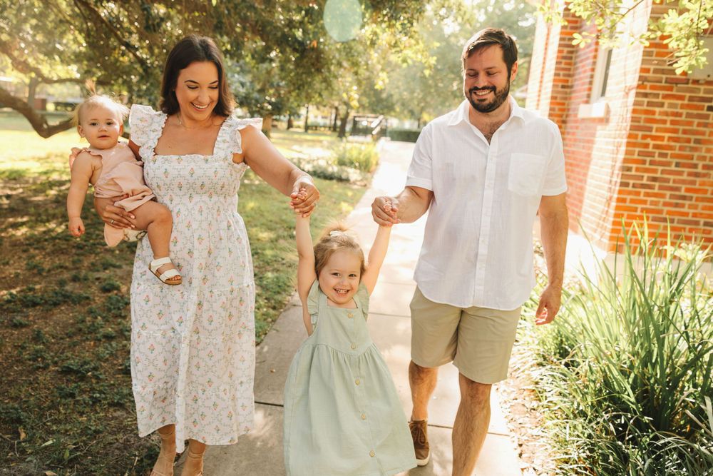 Happy young family of four walking outdoors, parents swinging toddler girl between them while holding baby.