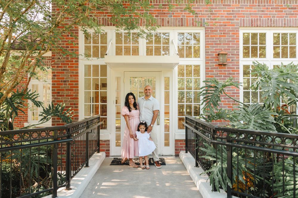 Family of three posing together on a bridge in front of a classic brick building.