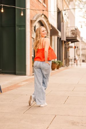 A senior girl in an orange top and jeans, looking over her shoulder, smiling on the urban sidewalk with string lights overhead. Huntsville