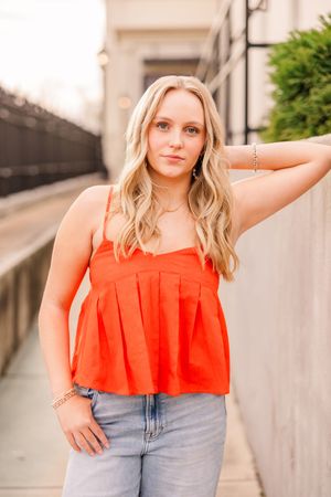 A senior girl in a bright orange pleated tank top and jeans is posing against a concrete wall outdoors.