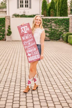 Person holding Texas A&M sign wearing denim skirt and cowboy boots on brick walkway.