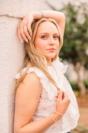 Portrait of person in white eyelet top leaning against light colored wall.