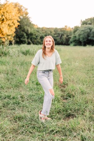 Young woman in casual attire laughing while standing in a sunlit meadow with autumn trees in background.